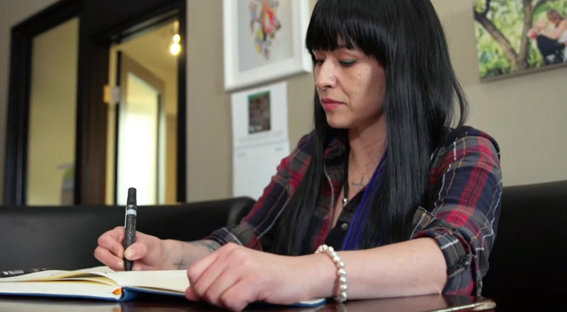 Woman seated at a table, writing in a notebook with a pen. Artwork and a wall calendar are visible mounted on a wall in the background.