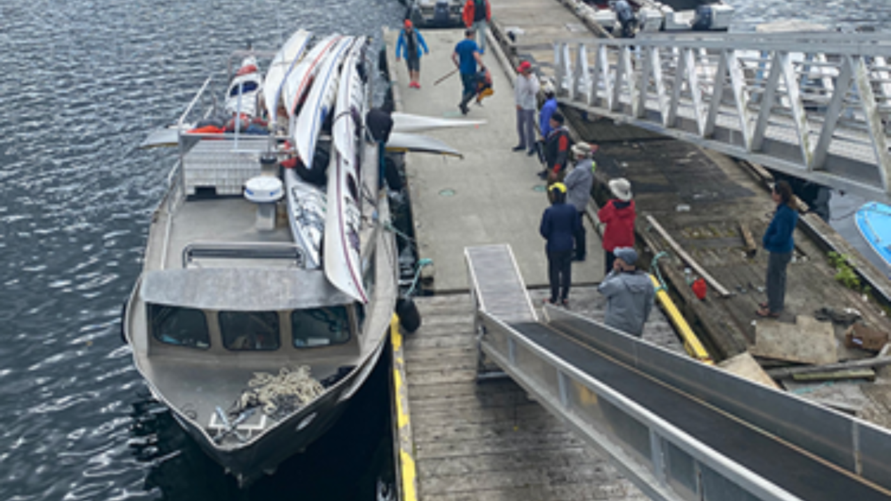 People standing on a dock in front of a YuWala Marine vessel with canoes attached to it