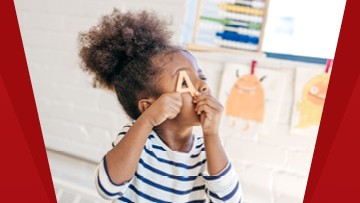 Une enfant tient la lettre « A », laquelle est faite de bois. Des dessins d’enfants sont affichés à l’arrière-plan.